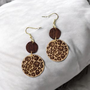 Könnte beinhalten: Ein Paar Ohrringe aus Holz mit Leopardenmuster. Die Ohrringe sind braun und goldfarben und haben einen schlichten, eleganten Stil.