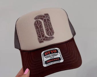 May include: A brown and beige trucker hat with a brown mesh back. The front of the hat features a brown graphic of two cowboy boots. The hat has a label that reads "OTTO WORLD FAMOUS FOAM FRONT OSFM TRUCKER DURABLE SNAP CLOSURE".