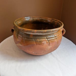 Puede incluir: Un cuenco de barro de color terracota con una abertura ancha, dos pequeñas asas laterales y un acabado vidriado. El exterior presenta una serie de líneas diagonales incisas alrededor de la sección superior, con variaciones en tonos marrones y bronceados.