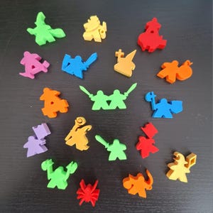 May include: A collection of colorful game pieces in various shapes, including figures with swords, shields, and other fantasy-themed designs. The pieces are in shades of green, yellow, red, blue, orange, and purple. The pieces are scattered on a dark surface.