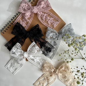 May include: Five different lace bows in various colors, including pink, black, gray, white, and beige. The bows are arranged on a white surface with a brown notebook in the background.