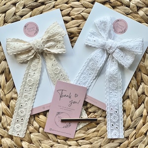 Könnte beinhalten: Zwei Haarschleifen aus Spitze, eine cremefarbene und eine weiße, mit floralen Lochdetails, auf weißen Karten präsentiert. Eine kleine rosa Karte mit dem Text "Thank you!" und eine goldene Haarspange sind ebenfalls zu sehen.