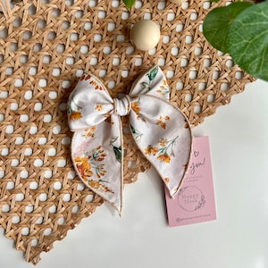 Könnte beinhalten: Ein weißes und gelbes Blumenhaarband mit cremefarbenem Satinbesatz. Die Schleife ist zu einem Knoten gebunden und liegt auf einer geflochtenen Weidenoberfläche. Der Text auf der rosa Karte lautet "Happy Nook" und "Vielen Dank! Ihr Kauf hilft zu unterstützen!"