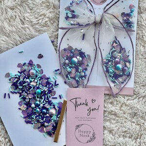 May include: A thank you card with a pink background and the text "Thank you!" in black. The card also features a small circle with the text "Happy Nook" in black. The card is surrounded by a pile of purple, blue, and white sprinkles.