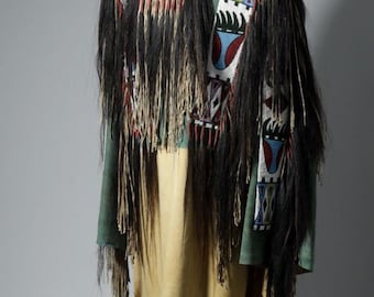 May include: A Native American ceremonial garment with a green top, tan bottom, and black fringe. The garment features colourful geometric beadwork and a black collar. The fringe hangs from the shoulders and sleeves.