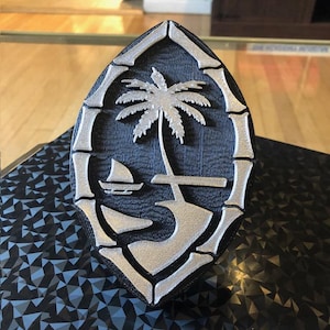 May include: A 3D printed silver and black shield with a palm tree and a sailboat on a beach. The shield is shaped like the coat of arms of Samoa.