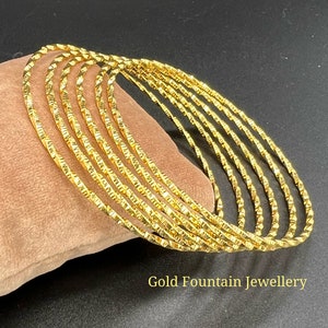May include: A set of gold bangles with a textured pattern, displayed on a beige cushion. The bangles are arranged in a circular formation, catching the light. The text "Gold Fountain Jewellery" is visible.