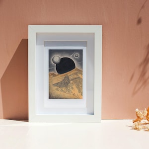 May include: A framed print of a black and white illustration depicting a landscape with a large black orb in the sky and a smaller white orb. The landscape is a desert scene with a mountain in the background.