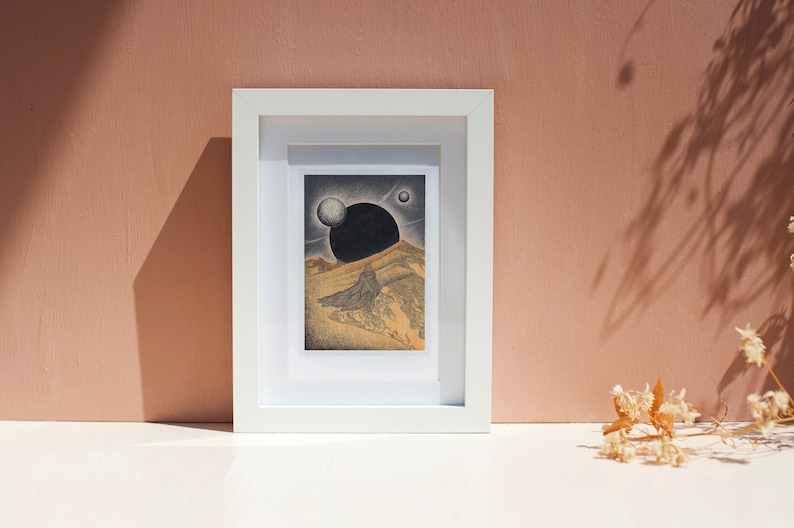 May include: A framed print of a black and white illustration depicting a landscape with a large black orb in the sky and a smaller white orb. The landscape is a desert scene with a mountain in the background.