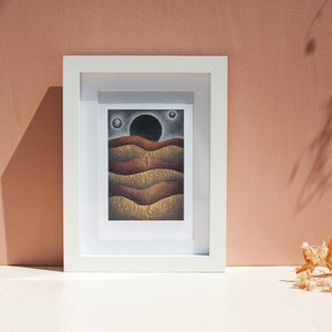 May include: A framed drawing of a landscape with a large black circle in the sky and a series of brown and gold hills.