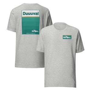 May include: Two heather grey t-shirts. One has a rectangular design on the back with the word "Duuuval" above a teal and gold striped pattern. The other shirt has a smaller matching logo on the front. The text "DUVAL CLOTHING CO" is below the design on the back.