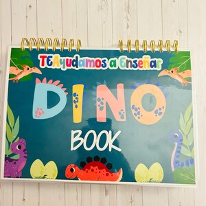 Puede incluir: Un libro de espiral azul verdoso con el título "DINO BOOK" en letras de colores. La portada presenta ilustraciones de dinosaurios, incluyendo un dinosaurio rojo con púas, un dinosaurio morado y un dinosaurio amarillo con cuello largo. El texto "TE Ayudamos a Enseñar" también es visible en la portada.