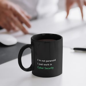 Op de afbeelding: Zwarte keramische mok met de tekst "I'm not paranoid I just work in Cyber Security" in wit en groen. De mok bevat een donkere vloeistof en staat op een wit oppervlak. Een computermuis en handen zijn op de achtergrond.