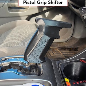 May include: A black pistol grip shifter installed in a vehicle. The shifter has a textured grip and is labelled "Pistol Grip Shifter" in a white box. The shifter is positioned near the gear selector and cup holders.