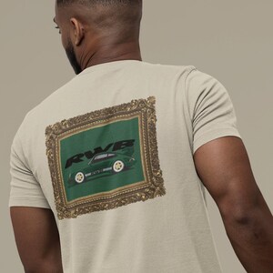 May include: Beige t-shirt featuring a framed graphic on the back. The graphic depicts a green sports car with the text "RWB" in black. The frame has a gold-colored ornate design. The t-shirt is short-sleeved.