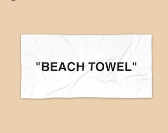 May include: White beach towel with the text "BEACH TOWEL" in black, centered on the towel. The towel is rectangular and appears to be made of a soft fabric. The background is a solid tan color.