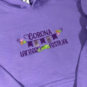 May include: A purple hoodie with embroidered text and graphics. The text reads "Corona Lantern Festival" in purple thread. The design includes a banner with lanterns, flowers, and a green chameleon.