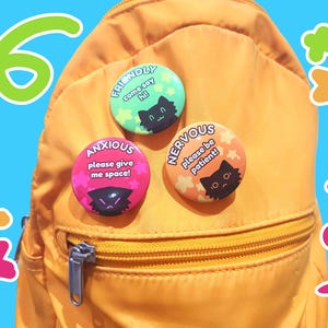 May include: Four colorful button pins with black cat illustrations and text that reads "Friendly come say hi!", "Anxious please give me space!", "Nervous please be patient!" and a pink, green, orange and yellow star design.