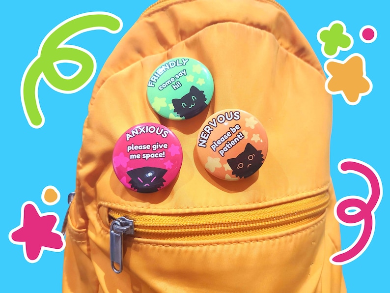 May include: Four colorful button pins with black cat illustrations and text that reads "Friendly come say hi!", "Anxious please give me space!", "Nervous please be patient!" and a pink, green, orange and yellow star design.