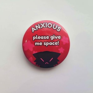 May include: Red button with white text that reads "ANXIOUS please give me space!" and a black cartoon character with a frown. Pink flowers surround the text.