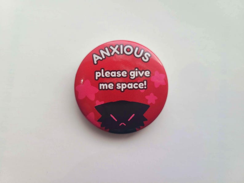 May include: Red button with white text that reads "ANXIOUS please give me space!" and a black cartoon character with a frown. Pink flowers surround the text.