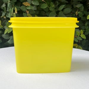 May include: A bright yellow, square-shaped container with a matching lid. The container is made of a solid, opaque material and sits on a white surface. The background features green foliage.