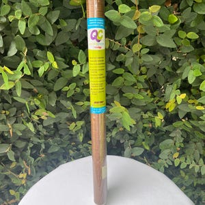 May include: A roll of brown adhesive covering with a wood grain pattern. The product is labeled "QC Quick Cover" and includes text about its features. The roll is displayed against a backdrop of green foliage.
