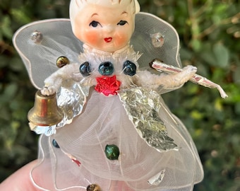 May include: A vintage-style fairy ornament featuring a ceramic head with painted facial features, white tulle wings, and a layered white tulle skirt. The figure is adorned with small metallic beads, silver foil accents, and holds a small gold-toned bell.