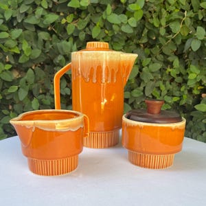 May include: A vintage orange ceramic coffee set with a creamer and sugar bowl. The coffee pot has a lid and handle. The set features a two-tone glaze with a drip effect. The sugar bowl has a wooden lid. The set is on a white surface with a green leafy background.