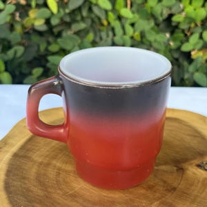 May include: A vintage coffee mug with a gradient design. The top of the mug is black, transitioning to red, with a white interior. The mug has a handle and sits on a wooden surface.