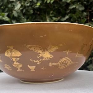 May include: A round, medium-brown mixing bowl featuring a repeating gold-colored pattern of an eagle, a lamp, a mortar and pestle, and a bellows. The bowl has a smooth, curved rim and a wide, deep basin, showcasing a classic mid-century design.