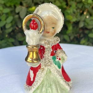 May include: A ceramic figurine of a person wearing a red coat with white glitter trim and a light green skirt. The figure wears a white glittery hat and holds a gold-colored candle holder with a red flame in front of their face.