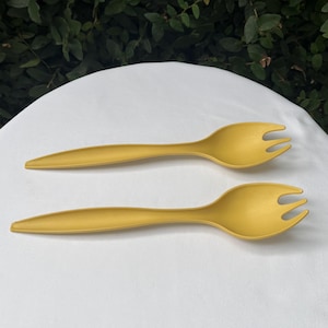 May include: Two mustard-yellow salad serving forks. Each fork has a long, slender handle and a three-tine head. The forks are made of a matte material and are set against a white background.