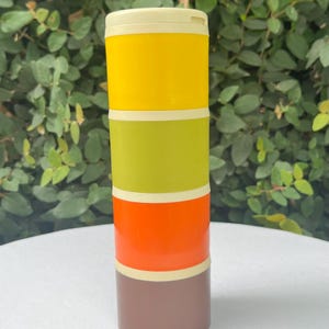 May include: A colorful, stacked container with a cream-colored lid. The container is divided into four sections: yellow, green, orange, and brown, each separated by a cream-colored band. The container is cylindrical and stands upright against a backdrop of green foliage.
