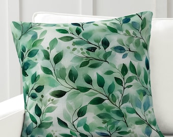 May include: A green and white throw pillow with a watercolor pattern of leaves and branches. The pillow is on a white couch.