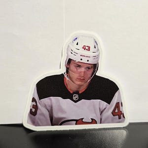 May include: A white hockey player with a white helmet with a red number 43 on it. The player is wearing a black and white jersey with a red logo. The player is looking down.