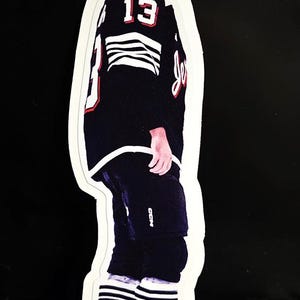 May include: A die-cut sticker of a hockey player in a black and red jersey with the number 13. The player is wearing a helmet and skates. The jersey has white stripes on the sleeves and the player's socks have white stripes.