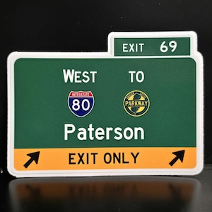 May include: A green and white sticker resembling a highway sign. It reads "WEST TO Paterson EXIT ONLY" with Interstate 80 and Garden State Parkway logos. The exit number is 69. Black arrows point to the right and left.