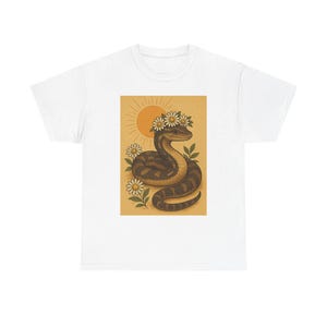 May include: White t-shirt with a graphic of a snake wearing a daisy crown, set against a yellow background with a sun illustration. The snake is brown and tan, with green leaves and white daisies. A casual, nature-themed tee.