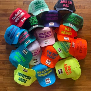 May include: A collection of colorful trucker hats featuring various phrases including Party Animal, Boat Day Bae, Too Glam To Give A Damn, Good Vibes Only, Old Enough To Know Better, Boat Hair Don't Care, Ship Show Supervisor, Drinks Well With Others, and Day Drunk.