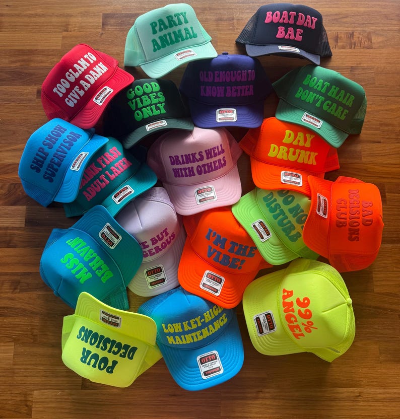 May include: A collection of colorful trucker hats featuring various phrases including Party Animal, Boat Day Bae, Too Glam To Give A Damn, Good Vibes Only, Old Enough To Know Better, Boat Hair Don't Care, Ship Show Supervisor, Drinks Well With Others, and Day Drunk.