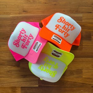 May include: Three trucker hats with white fronts and mesh backs. The hats are in bright colors: pink, orange, and neon yellow. Each hat has the phrase "Shorty hit Forty" printed on the front in a matching color.