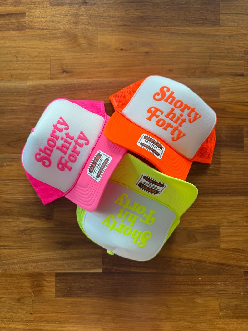May include: Three trucker hats with white fronts and mesh backs. The hats are in bright colors: pink, orange, and neon yellow. Each hat has the phrase "Shorty hit Forty" printed on the front in a matching color.