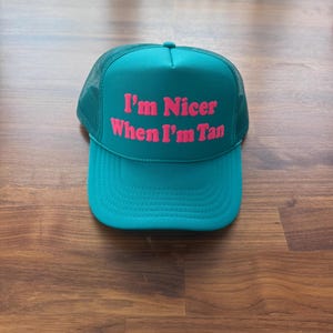 May include: A teal trucker hat featuring a mesh back and a solid front panel. The text "I'm Nicer When I'm Tan" is printed in bold, bright pink lettering across the center of the front panel. The hat has a curved brim and a structured crown.
