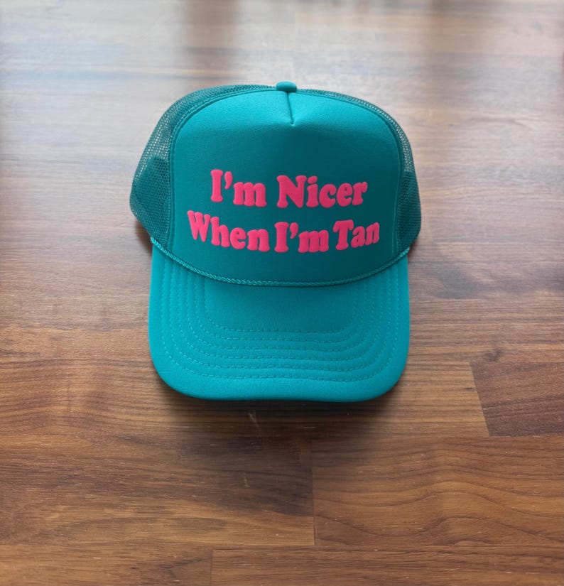May include: A teal trucker hat featuring a mesh back and a solid front panel. The text "I'm Nicer When I'm Tan" is printed in bold, bright pink lettering across the center of the front panel. The hat has a curved brim and a structured crown.