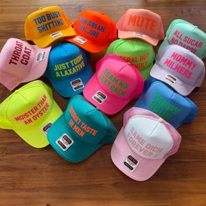 May include: A collection of twelve colorful foam trucker hats featuring various phrases in bold lettering. The hats come in shades of yellow, orange, pink, blue, green, and white, each with a structured front panel and mesh back, displaying text like Mute, Yappy Hour, and Throat Coat.