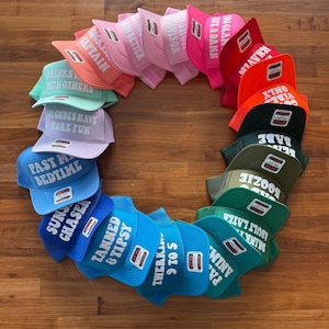 May include: A collection of colorful trucker hats arranged in a circle, each featuring white text including phrases like 'Tanned & Tipsy', 'Past My Bedtime', 'Blondes Have More Fun', 'Drinks Well With Others', 'Cocktail Captain', and '9 To 5'.