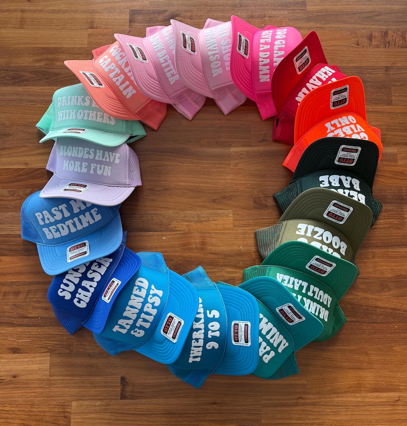 May include: A collection of colorful trucker hats arranged in a circle, each featuring white text including phrases like 'Tanned & Tipsy', 'Past My Bedtime', 'Blondes Have More Fun', 'Drinks Well With Others', 'Cocktail Captain', and '9 To 5'.