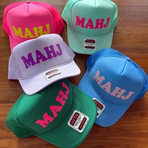 May include: A collection of trucker hats in various colors, including pink, green, blue, and purple. Each hat features the word "MAHJ" in a contrasting color across the front. The hats have a mesh back and a curved brim.