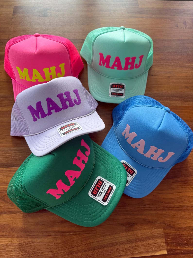 May include: A collection of trucker hats in various colors, including pink, green, blue, and purple. Each hat features the word "MAHJ" in a contrasting color across the front. The hats have a mesh back and a curved brim.